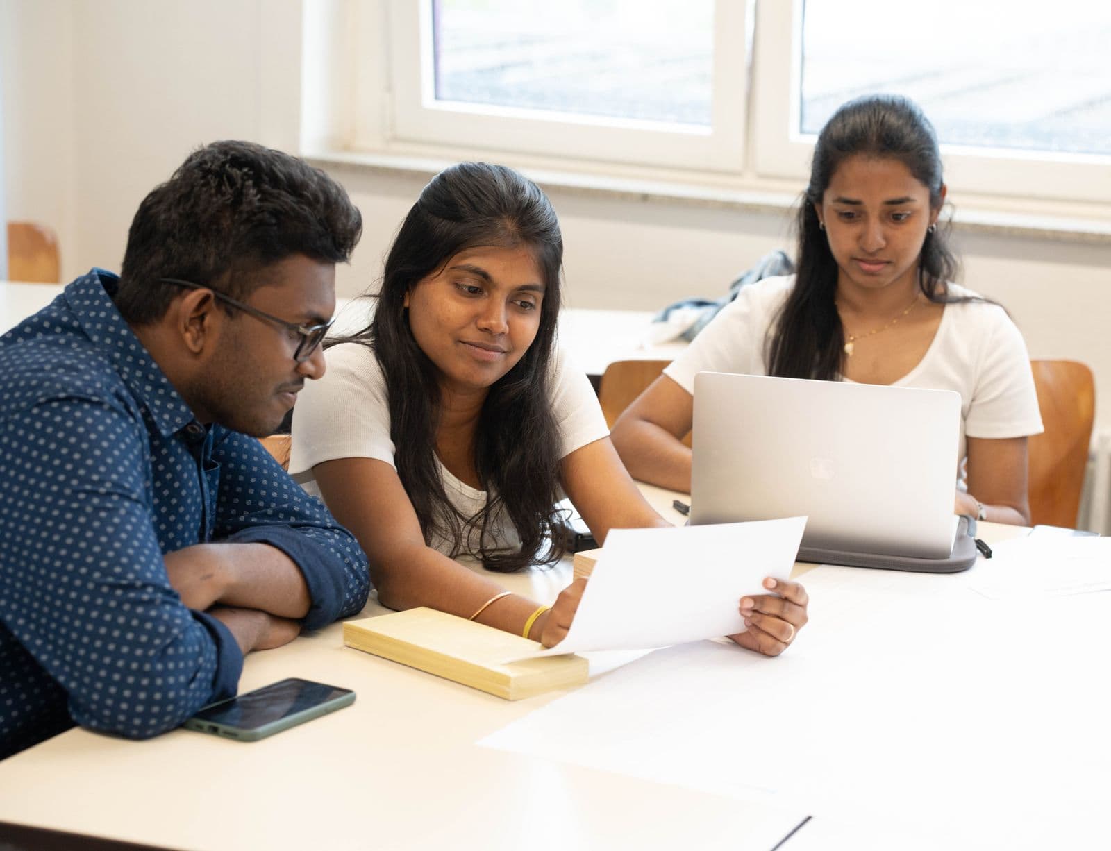 Three South Asian students, two of them women, studying together over a laptop in a bright classroom