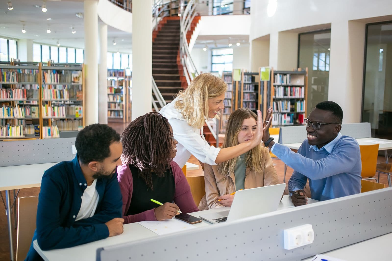 A diverse group of learners — Black, white and mixed-heritage, women and men — celebrating progress together over a laptop in a modern library